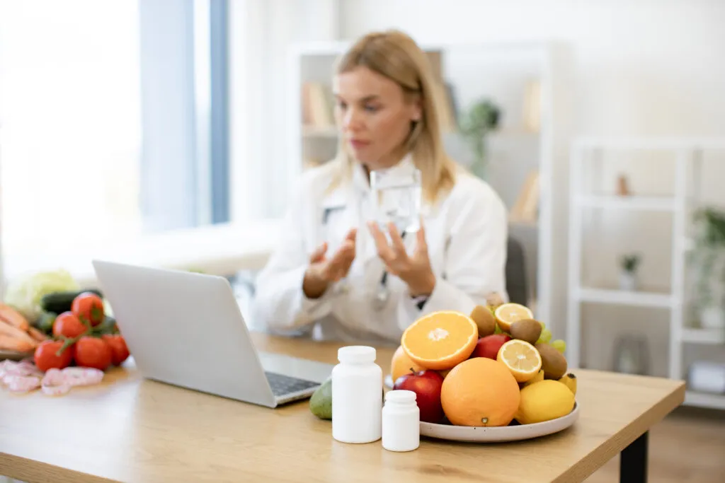 Nutricionista analisando frascos de vitaminas ao lado de uma tigela com frutas variadas, incluindo laranjas, maçãs, uvas e abacate, em ambiente bem iluminado.