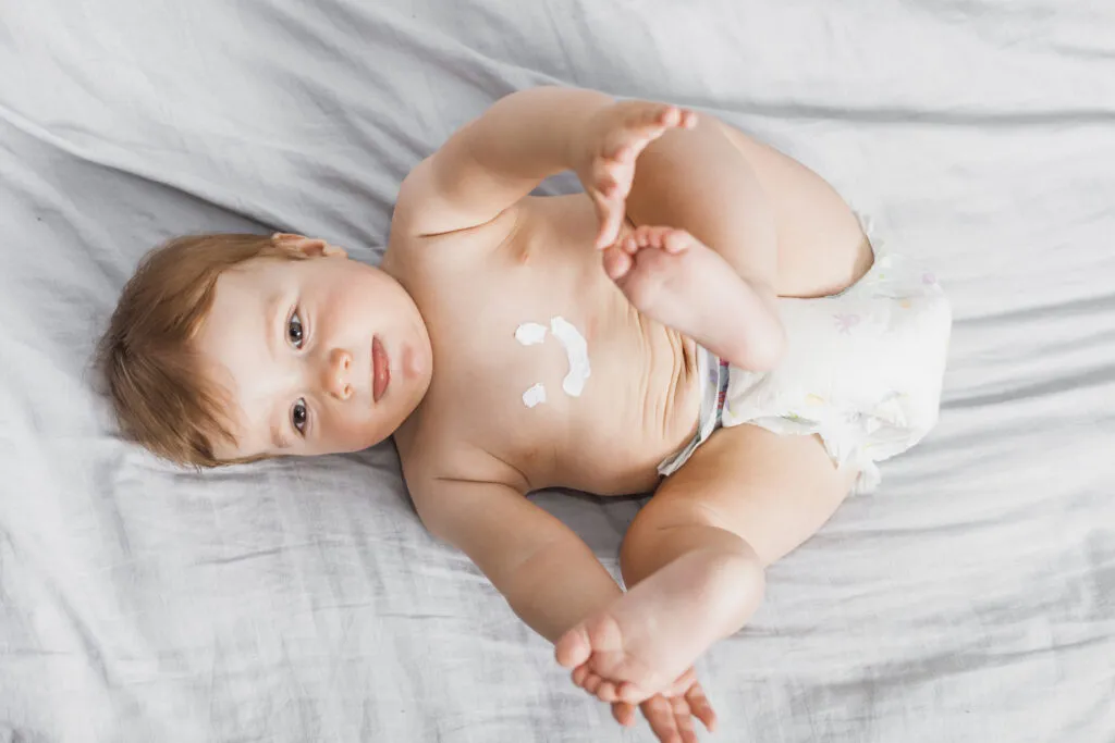 Bebê deitado em berço, exibindo expressiva curiosidade e com pequenas manchas de leite na barriga, em um ambiente confortável e acolhedor.