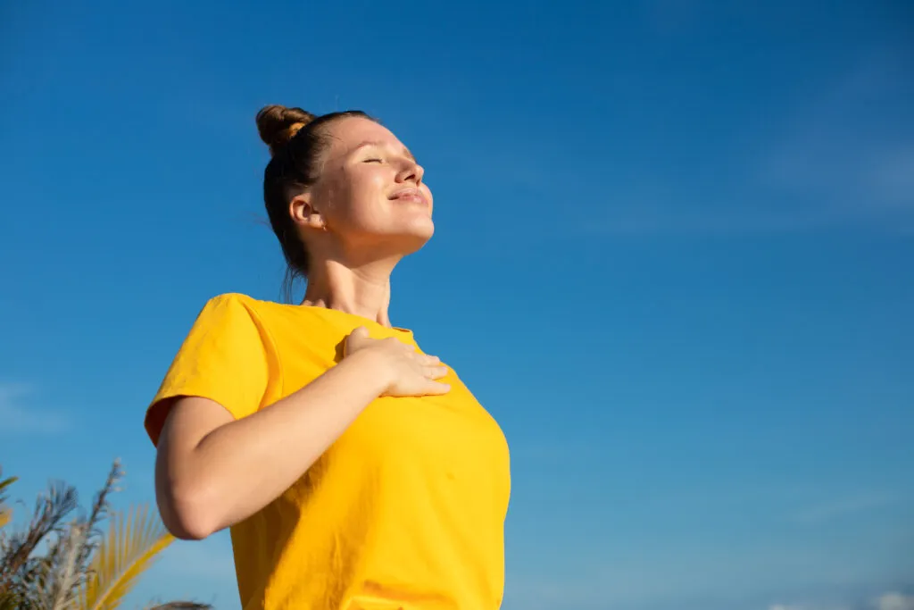 Mulher sorridente com expressão de paz e gratidão, usando camiseta amarela, aprecia o ar livre ao sol com um céu azul ao fundo.