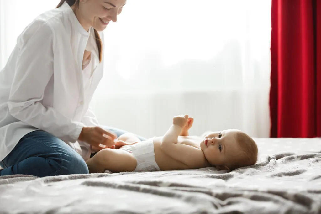 Mãe brincando com seu bebê deitado na cama, sorrindo e se divertindo em um ambiente acolhedor e iluminado, com cortinas vermelhas ao fundo.