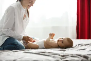 Mãe brincando com seu bebê deitado na cama, sorrindo e se divertindo em um ambiente acolhedor e iluminado, com cortinas vermelhas ao fundo.
