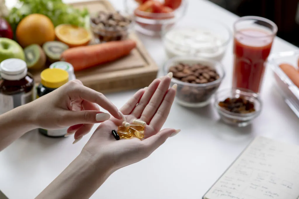 Imagem de mãos segurando cápsulas de suplemento alimentar em um ambiente com alimentos saudáveis como frutas e vegetais ao fundo, promovendo nutrição e saúde.