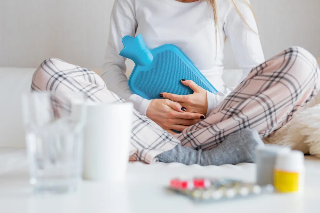 Pessoa com dor menstrual segurando bolsa de água quente e remédios, sentada no sofá com roupas confortáveis, mostrando alívio na região abdominal.