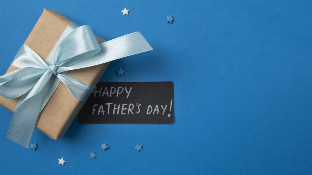 Presente embrulhado com laço azul e cartão preto com mensagem “HAPPY FATHER’S DAY” sobre fundo azul, com pequenos enfeites brilhantes ao redor, ideal para Dia dos Pais.