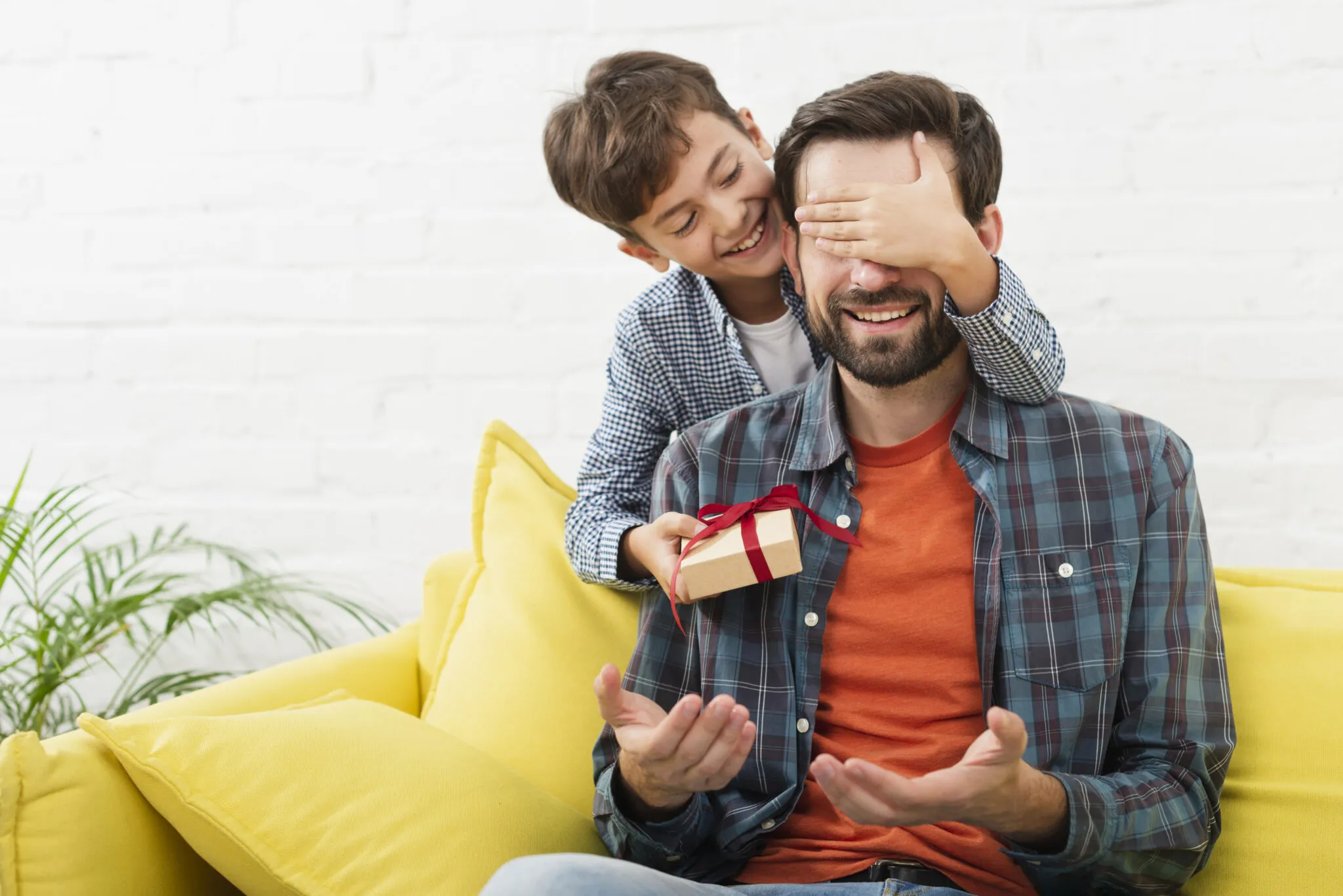 Pai sorrindo no sofá enquanto o filho deita a mão sobre os olhos dele e brinca de surpresa, segurando um pequeno presente com fita vermelha.