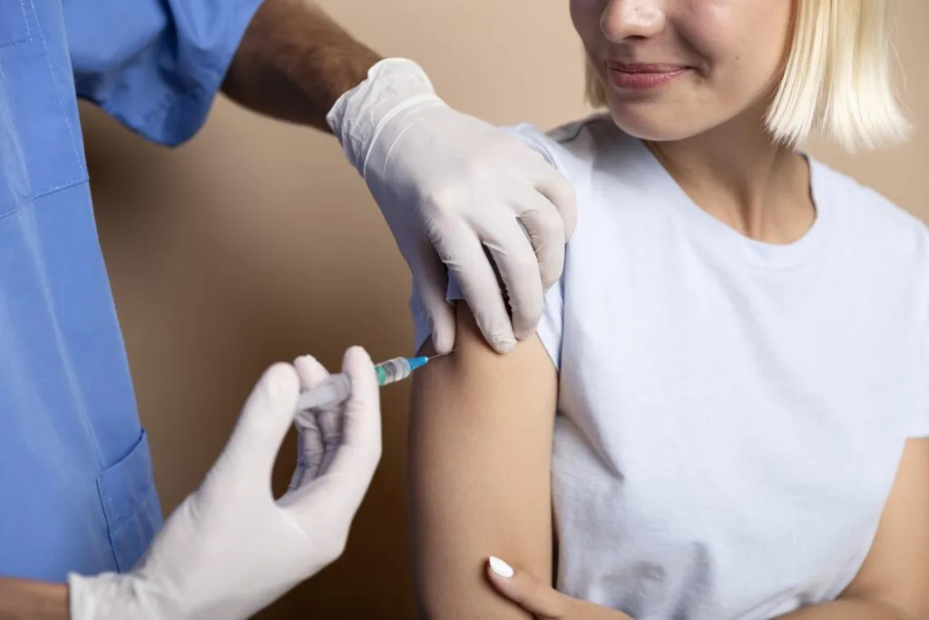 Profissional de saúde aplicando vacina no braço de uma mulher, com seringa e algodão esterilizado, em ambiente clínico, mostrando o cuidado durante a imunização.